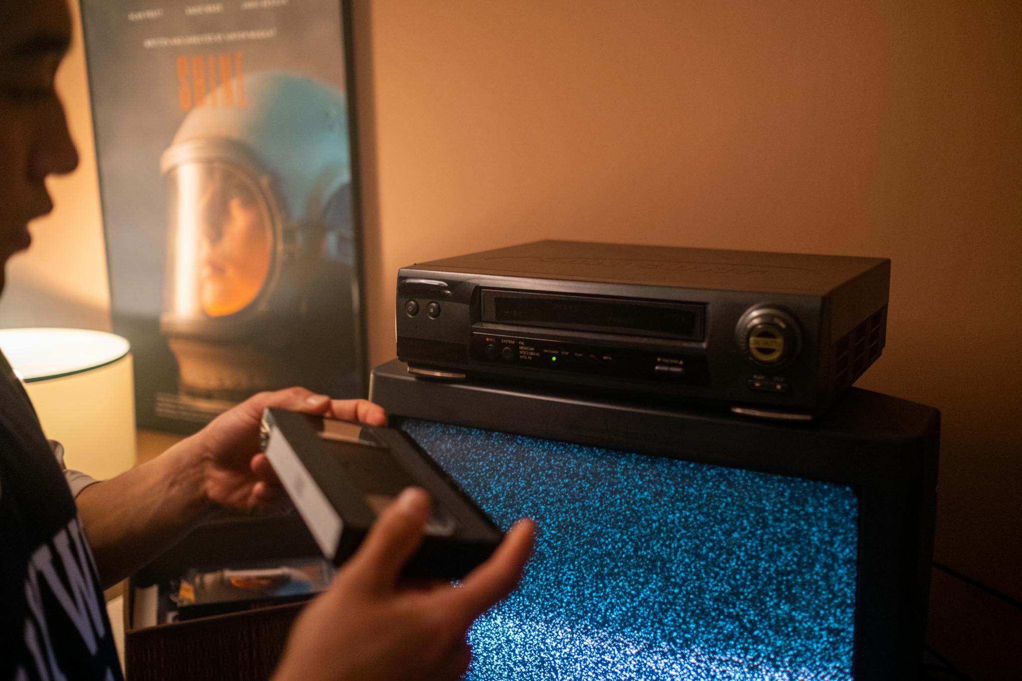 Person inserting VHS tape into video recorder with static TV in cozy room.